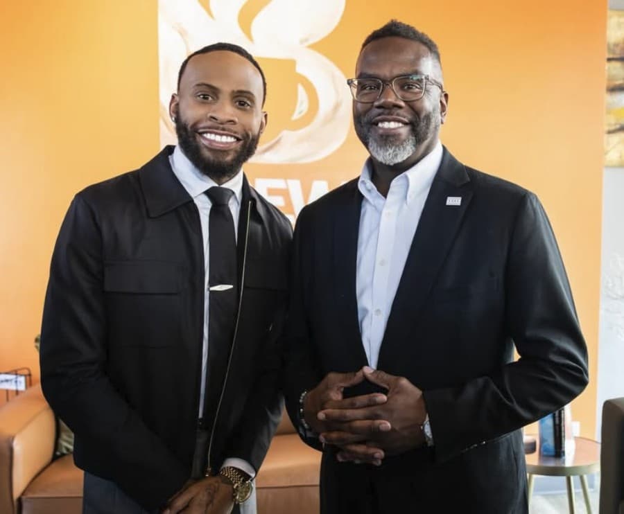 Two men in dark jackets smiling in front of The Brew's orange logo wall